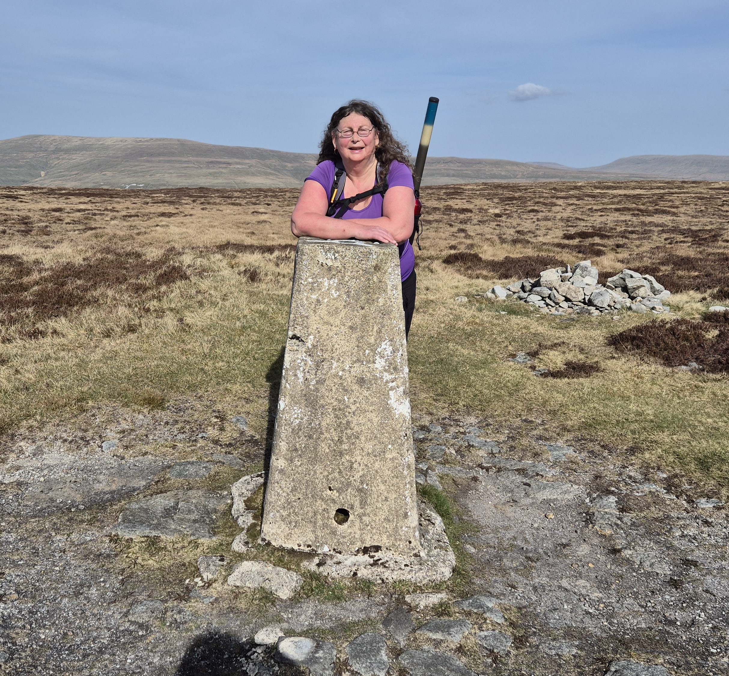 Carolyn is wearing a purple top and leaning on a trig point